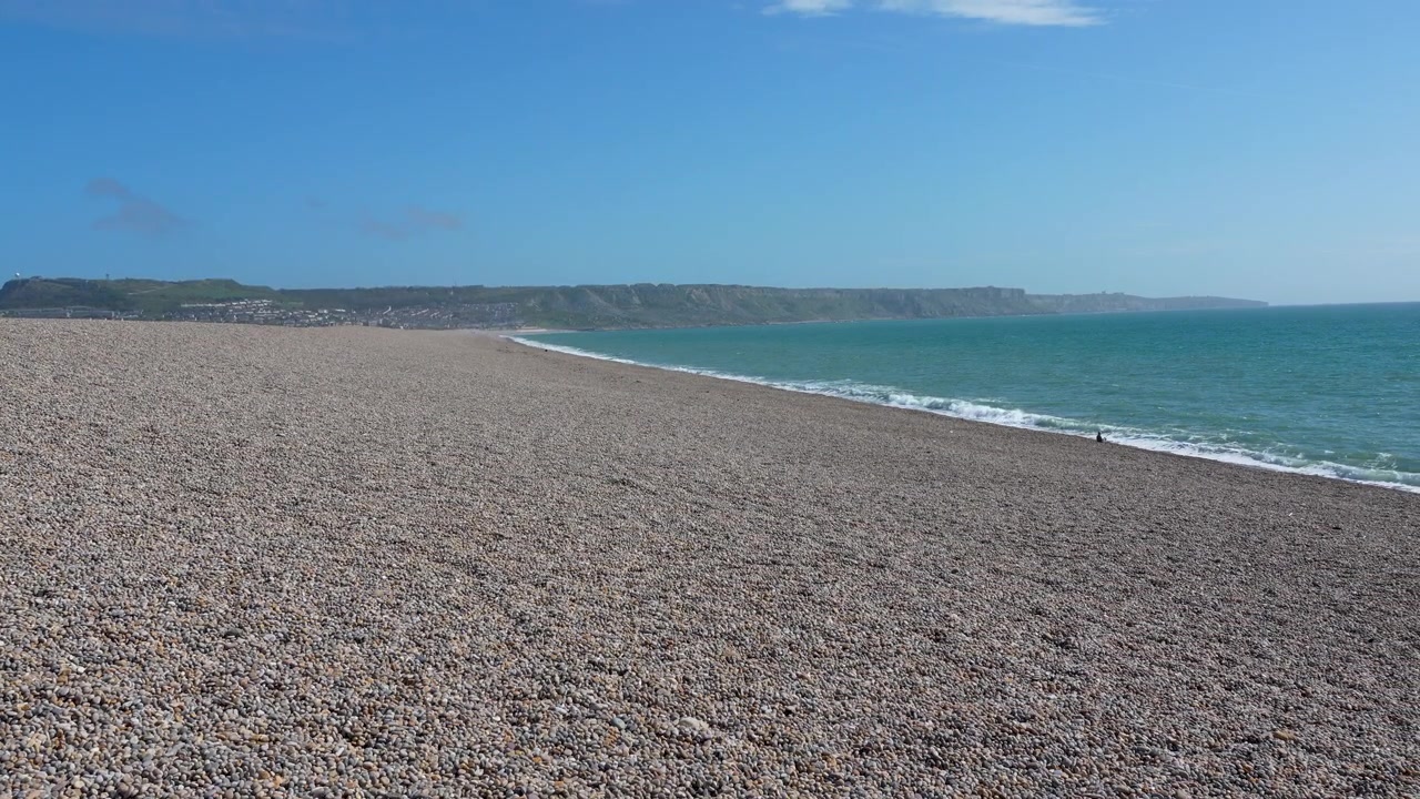 Un'ampia inquadratura panoramica dell'iconica costa di ciottoli a Chesil Beach a Weymouth, con le scogliere della Jurassic Coast sotto un cielo azzurro e limpido. Ideale per racconti di viaggio e ambientali