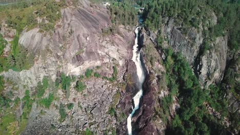 Backwards-tilt-down-drone-shot-of-a-large-waterfall-in-Norway-and-a-green-forest-in-the-background