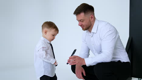 proud father in white shirt and his son in white shirt and black tie. father shows a smartphone to his baby boy.
