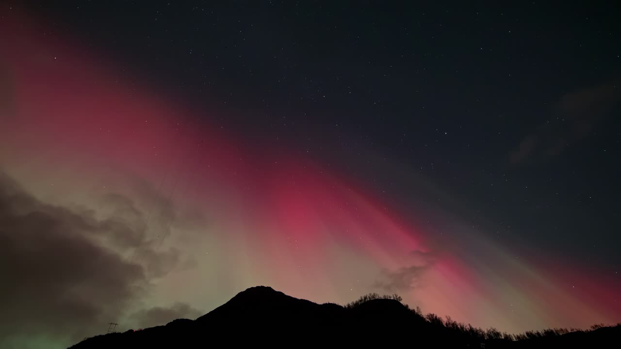 Stunning northern lights over Norway with green and red aurora dancing across the night sky. Moving clouds and dark mountain silhouette below