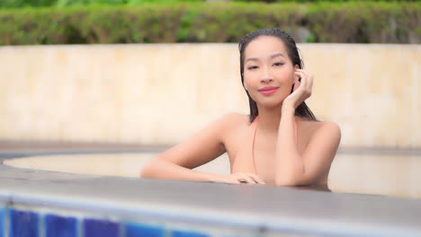 face closeup asian woman in water in the swimming pool, she has wet hair, smiling and turning her sight towards camera