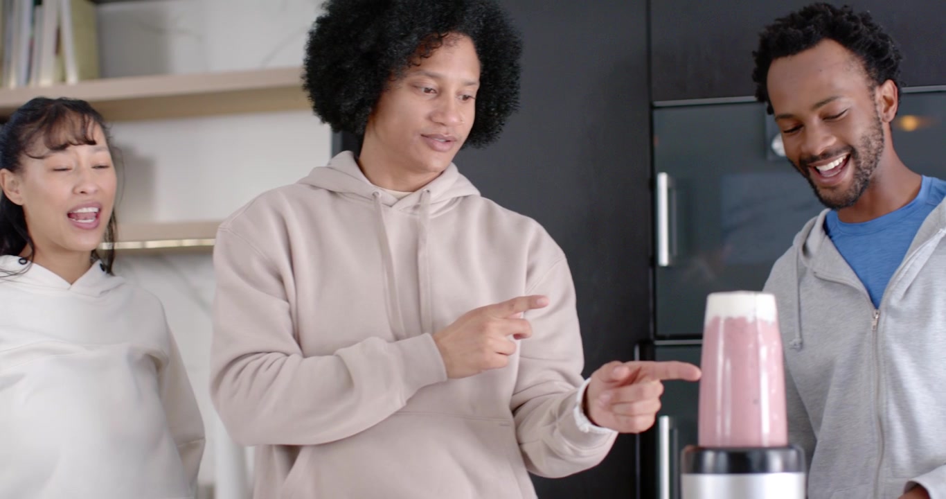 Diverse friends placing hand on blender lid and pointing at pink smoothie, checking foam in kitchen