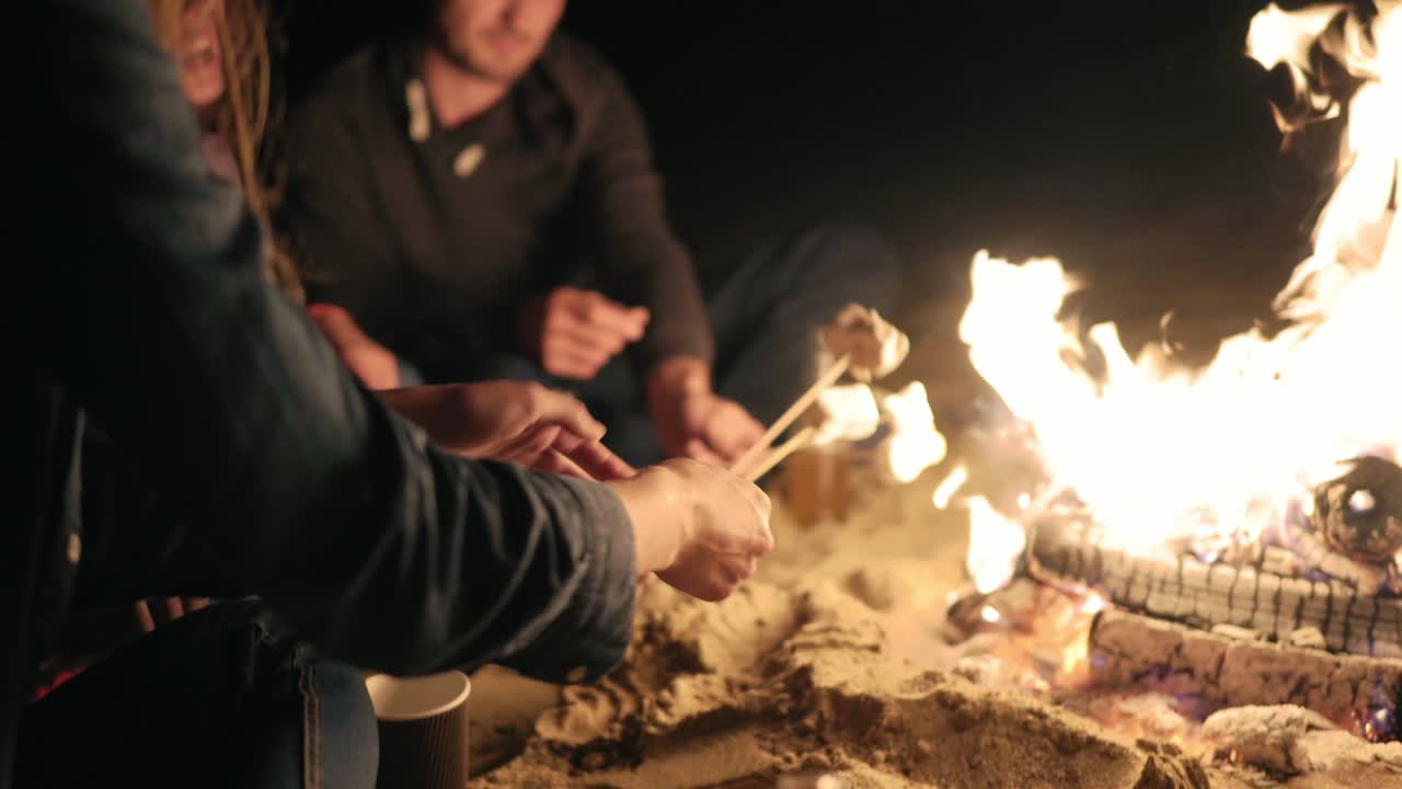 группа молодых многоэтнических людей, сидящих у огня на пляже поздно ночью, жареные зефиры на палках над огнем вместе