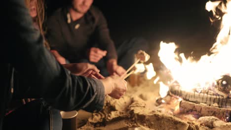 Grupo-De-Jóvenes-Multiétnicos-Sentados-Junto-Al-Fuego-En-La-Playa-A-Altas-Horas-De-La-Noche,-Asando-Juntos-Malvaviscos-En-Palos-Sobre-El-Fuego