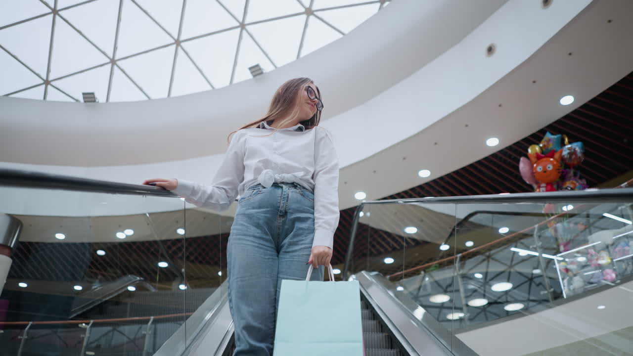 Ung kvinna som håller i shoppingkassar kliver på en rulltrappa nedåt i ett modernt köpcentrum, lägger handen på räcket medan hon tittar bortåt, omgiven av starkt ljus