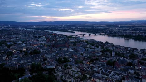 Revealing-Drone-shot-of-Mainz-at-magic-hour-night-showing-a-colorful-sky-and-reflections-of-the-Rhine-river-water-in-the-background