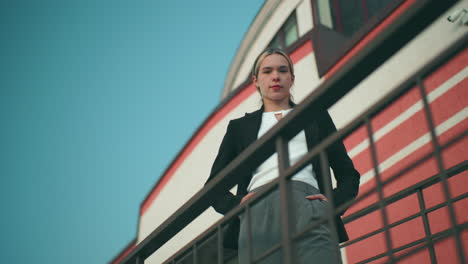 upward view of confident lady posing with warm smile near iron railing with striped office building in background under clear blue sky, hands in pockets and radiating poise