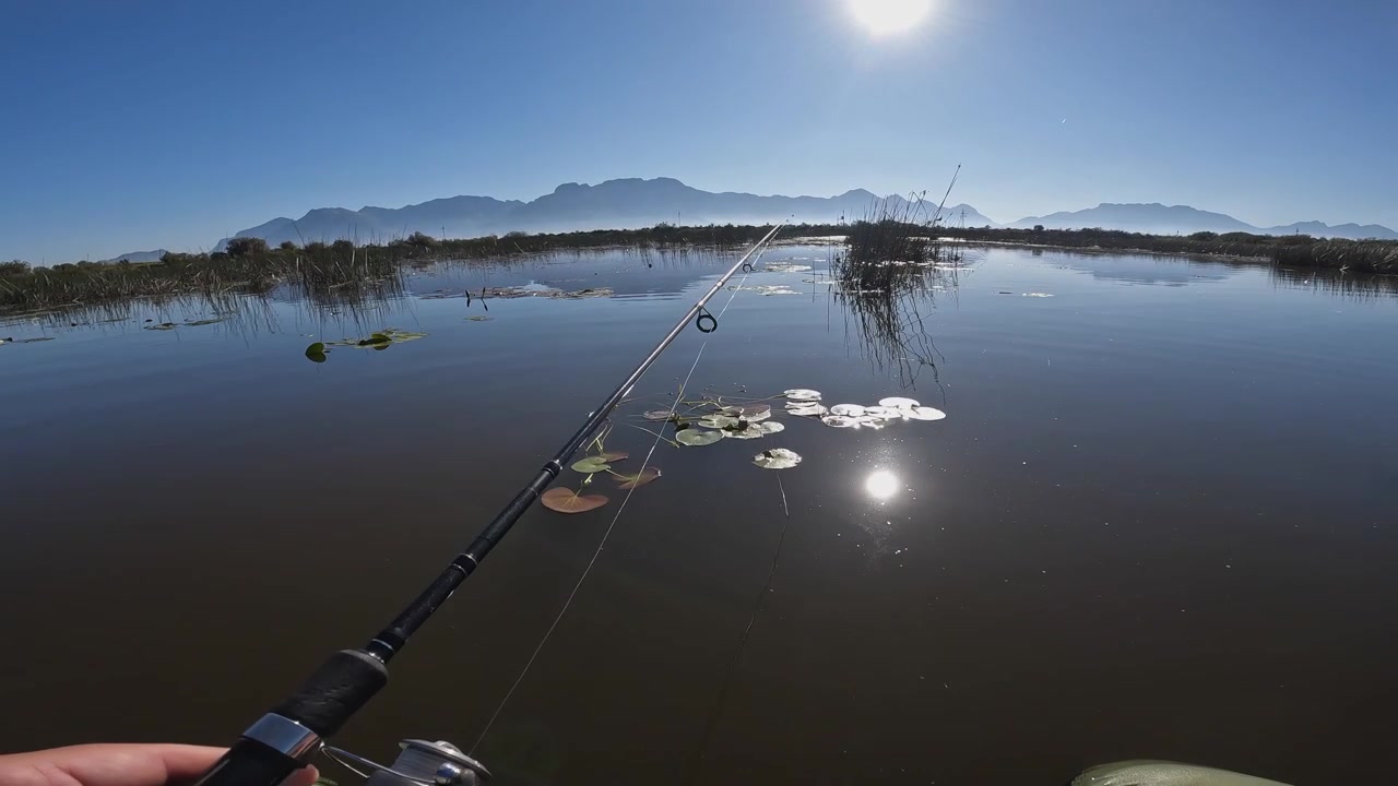 Man fishing POV in blue lake, morning sun shines over distant mountains