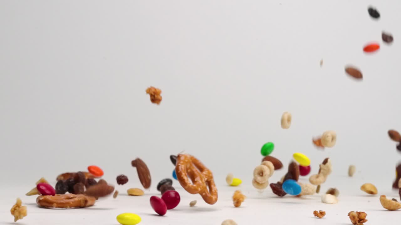Sweet and salty trail snack mix with peanuts, pecan, almond, and cashew nuts, pretzels, m and m candies and cheerios falling onto white table top in slow mo