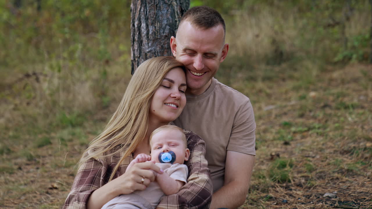 Relaxed blonde long-haired woman leaned on her husband sitting behind. Couple with baby spend time in the nature.
