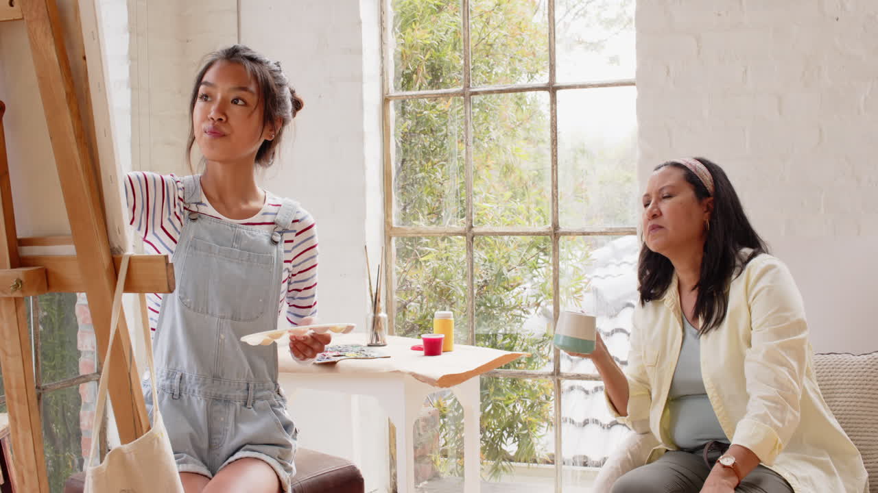 Multiracial young woman painting while grandmother enjoys coffee, sharing creative moment at home