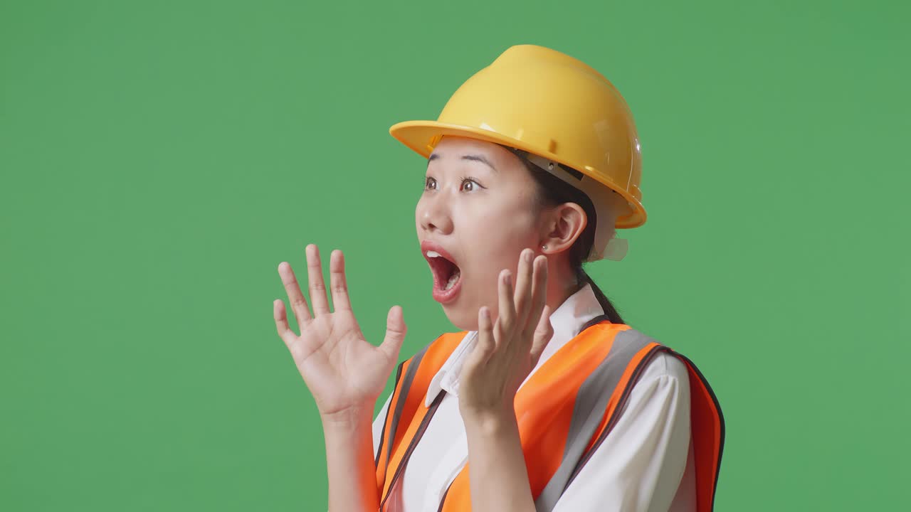 안전 헬을 입은 아시아 여성 엔지니어가 녹색 스크린 배경 스튜디오에 서서 웃으며 와우라고 말합니다.