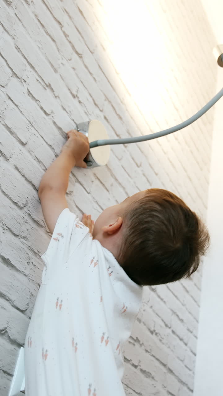 Little cute boy stands holding by the white brick wall. Curious toddler reaches the lamp above him to switch it off and on again. Low angle view. Vertical video