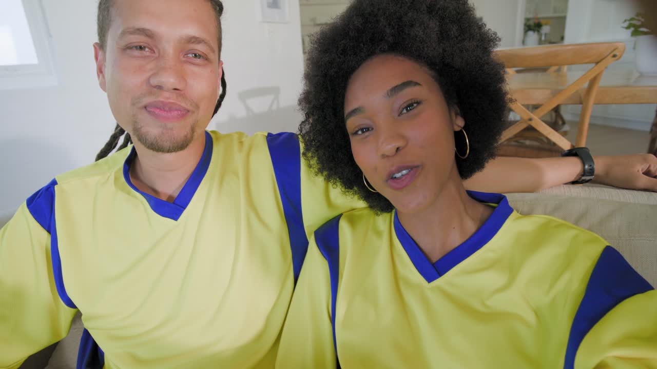 Amici afroamericani seduti sul divano con le maglie sportive, un uomo in grigio che si gira, una donna che filma un selfie con il telefono.