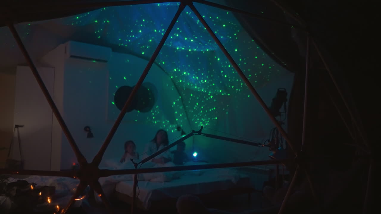 los niños se entretienen con la madre en bata de baño en una habitación débilmente iluminada. casa de brillo transparente con cielo nocturno estrellado en el techo iluminado por un proyector contemporáneo