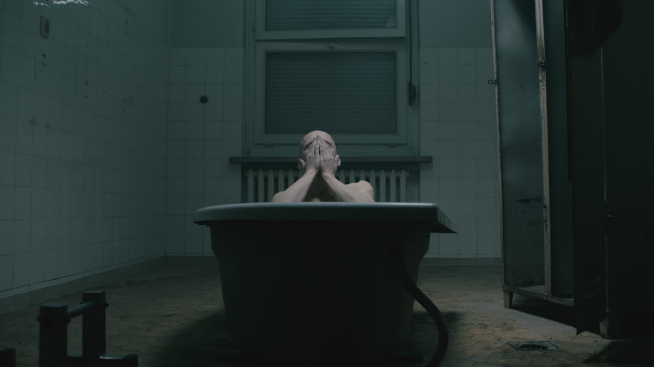 Pale bald skinny man takes a bath in the creepy old bathroom at night