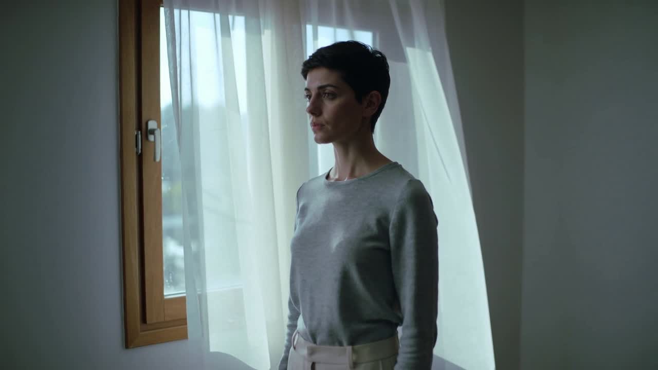 Donna in piedi che guarda vicino a una finestra di legno, la brezza muove la tenda velata, con un top grigio e pantaloni con cintura