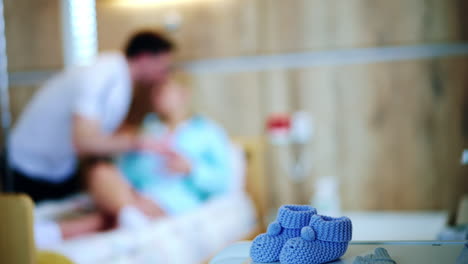 little blue booties waiting for the newborn. close up. cute socks on the table in hospital ward. couple at backdrop in blur.
