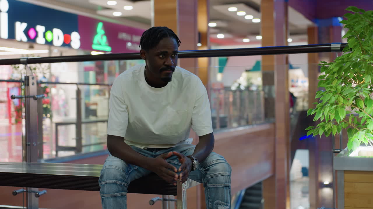 Man wearing white shirt and jeans seated on bench in mall interior tapping hands together with thoughtful expression modern retail lifestyle environment colorful blurred background