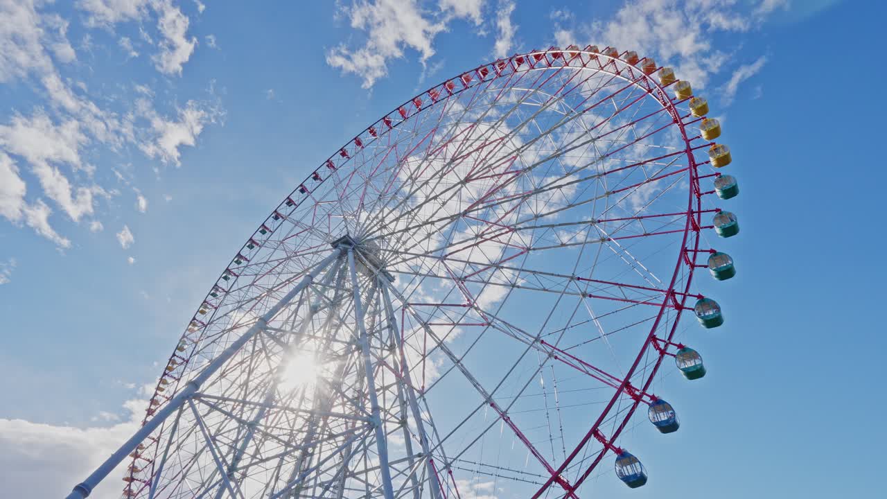 Dramatic low-angle view of a giant white and red Ferris wheel, with the sun flaring brightly from behind its central structure