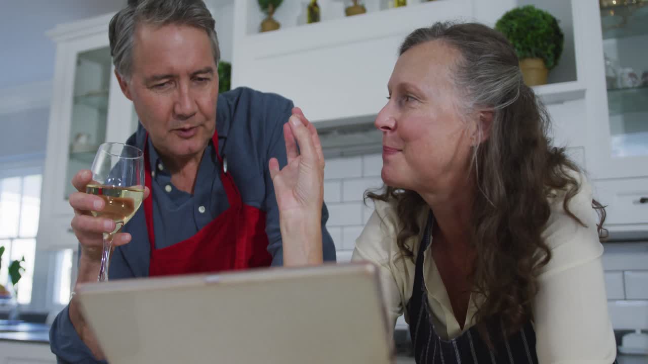 feliz pareja caucásica mayor en la cocina mirando la computadora portátil, hablando y disfrutando de un vaso de vino blanco