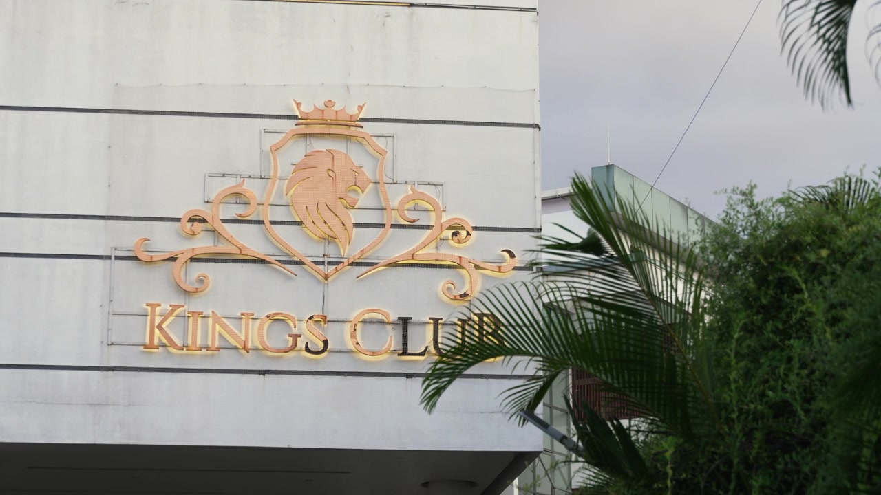 Primo piano dell'insegna d'ingresso di un Luxury Sports Club con stemma di leone dorato su una parete esterna bianca parzialmente incorniciata da fogliame verde.