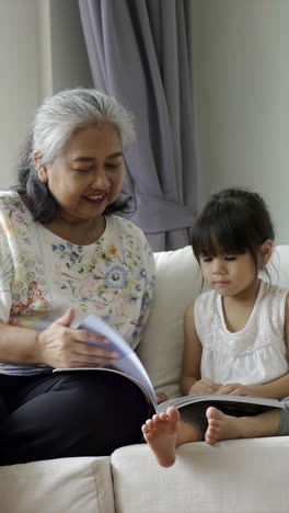 senior lifestyle concept: an affectionate asian grandmother patiently teaches her granddaughter to read and write while sitting on a sofa in the cozy comfort of their living room