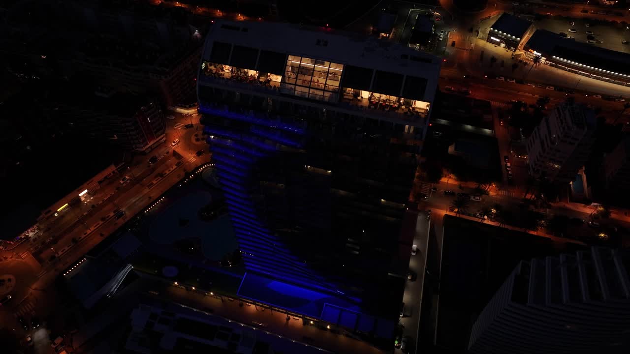 Night drone orbit captures Estimar Hotel in Calp , Spain, with its distinctive curved architecture, rooftop lounge, and deep blue light reflections, surrounded by illuminated city streets, slow motion