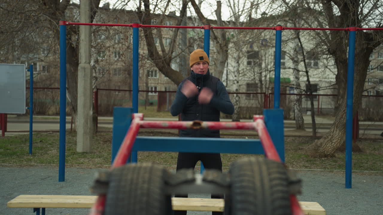 Close-up of a coach working out in an outdoor gym, the coach is looks to the left, and a dog is seen walking in the background across a road, with red and blue iron bar around
