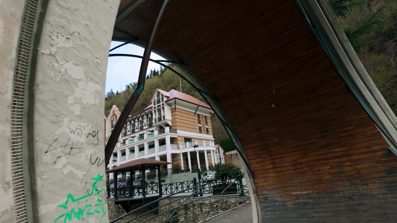 Ponte coperto architettonicamente suggestivo con un design curvo attraversa un fiume a Borjomi, Georgia, conducendo verso un edificio caratteristico annidato sul fianco della montagna. Sentiero pedonale attraverso un paesaggio scenografico naturale.