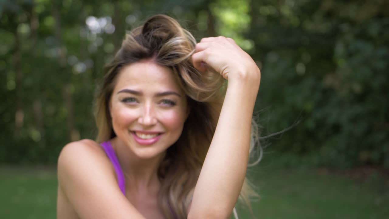una mujer bonita con cabello rubio usando ropa para hacer ejercicio en un parque sonríe y tira su cabello mientras sopla una suave brisa