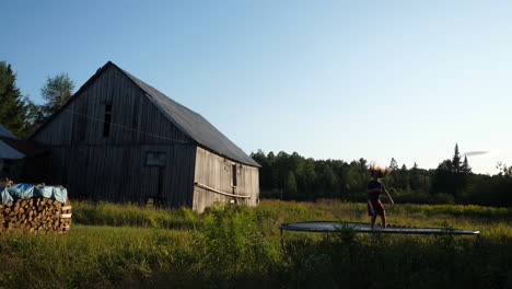 kid using a trampoline at a farm with wood stack, barn, forest and field