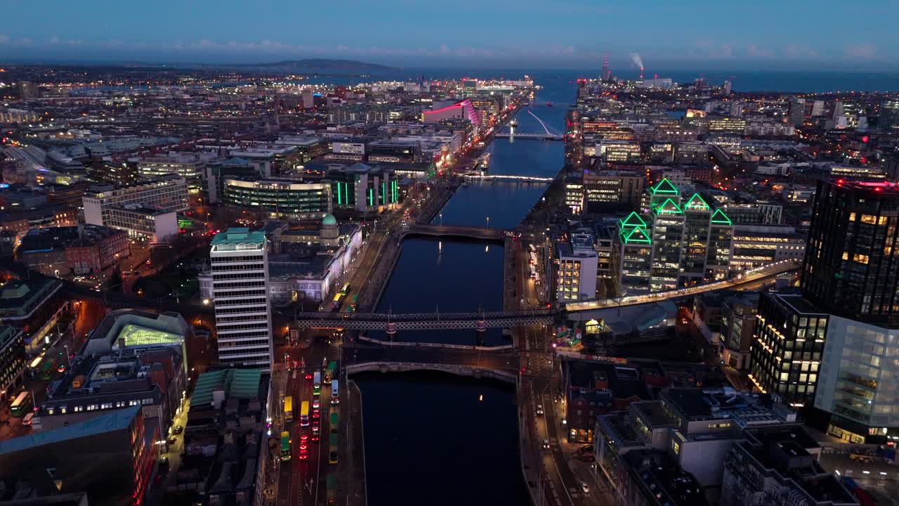 Luftaufnahme einer Drohne, die im Winter in der blauen Stunde der Dämmerung über den Fluss Liffey in Dublin fliegt, mit irischen, beleuchteten Stadtgebäuden und Brücken über dem Wasser, belebten Straßen und TouristInnen