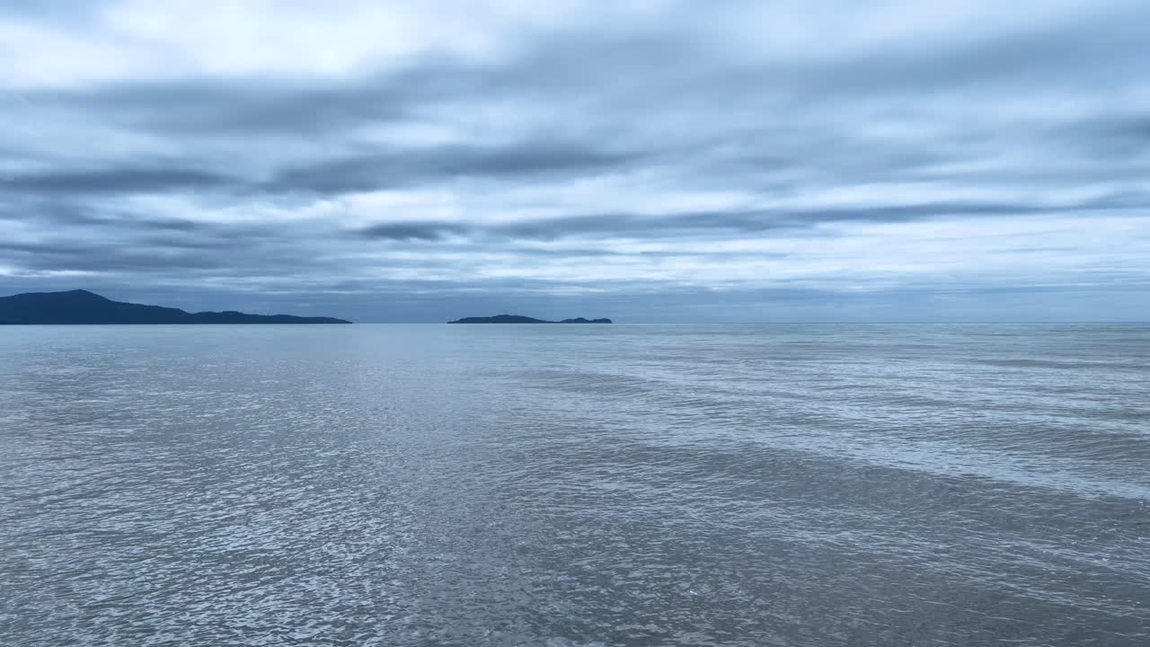 Camera slowly pans across calm ocean, rocky shore, distant islands, under dramatic cloudy sky