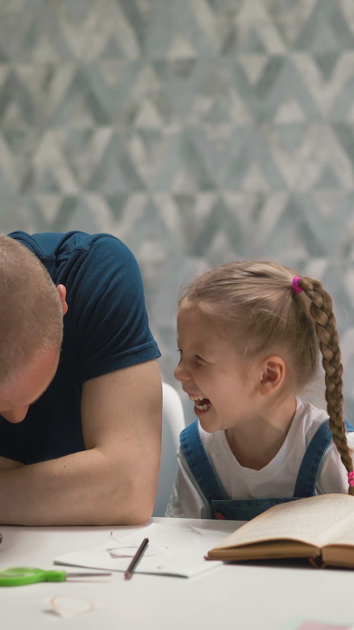 bald father working on laptop and pretty little daughter doing homework laugh sitting at white table in brightly lit room at quarantine time