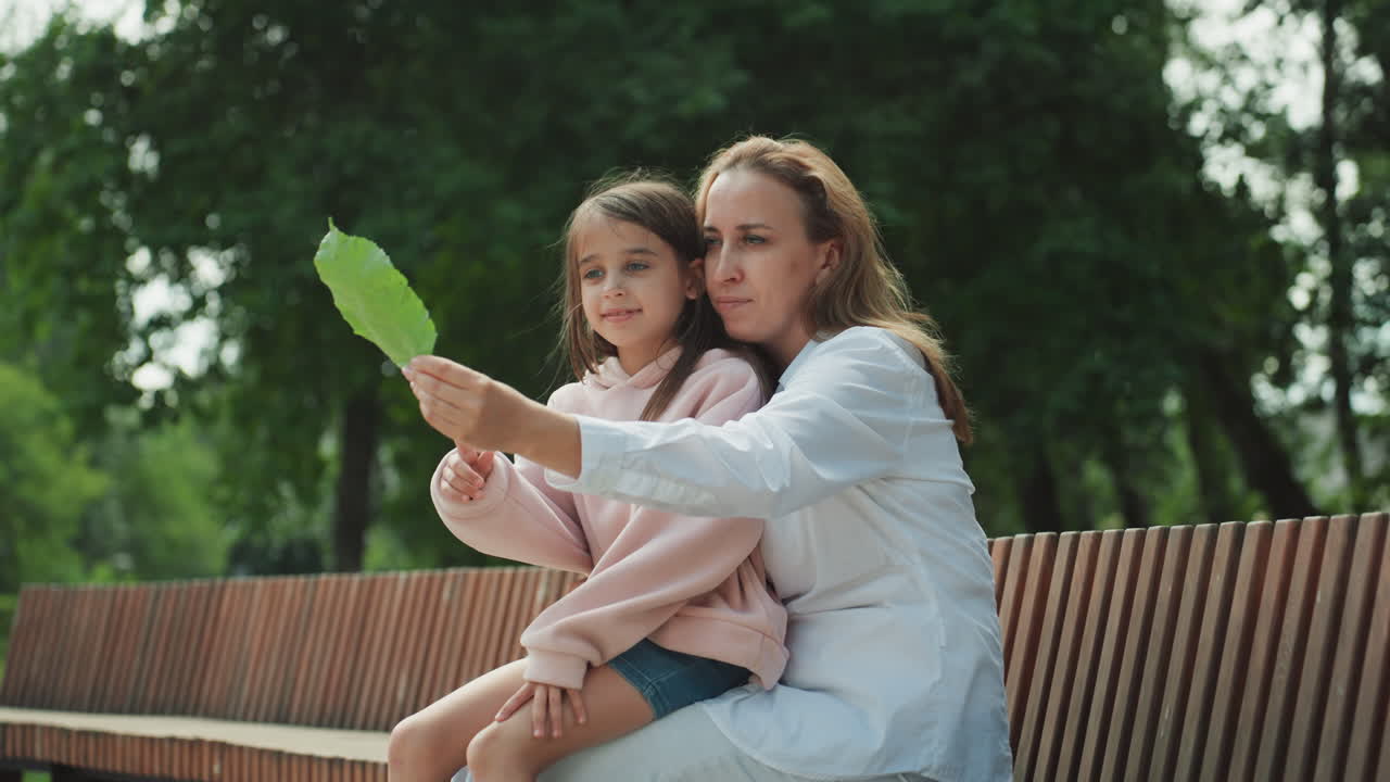 Mamma e figlia su una panchina del parco esaminano una foglia, la madre guida la bambina in una delicata lezione sulla natura con espressioni attente, sfondo di fogliame verde, luce calda del giorno, abbigliamento comodo, interazione delicata.