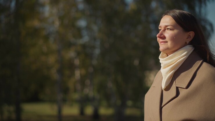Close up of woman wearing brown coat and cream scarf standing outdoors in natural light. Her hair is long dark and loose with small earrings visible. background features blurred trees