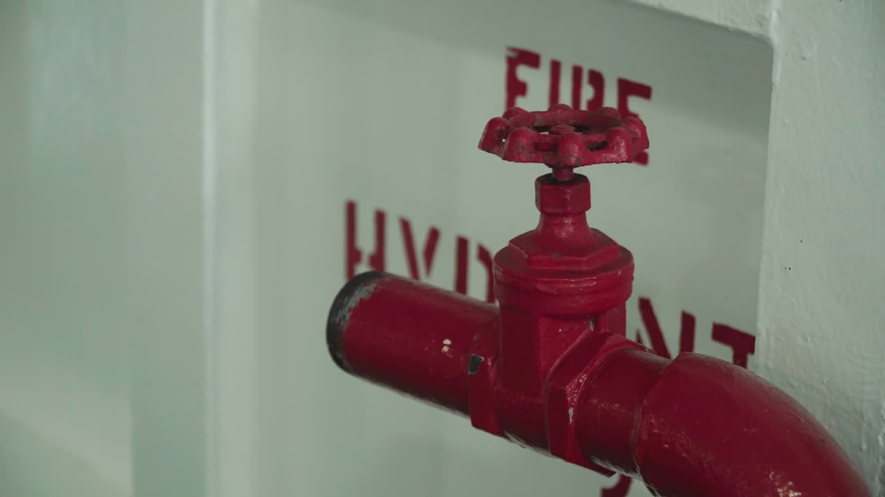 Valvola antincendio rossa contro il muro in un corridoio di servizio, primo piano di un tubo metallico verniciato e di una manopola a volantino, segnaletica di emergenza visibile sul pannello, la scena suggerisce un'ispezione di manutenzione e la conformità alle norme di sicurezza