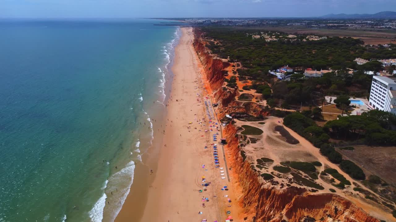Drone footage of Praia da Falesia shows long sandy beach bordered by red cliffs and turquoise sea with sunbathers and umbrellas under clear sky in Algarve region of southern Portugal