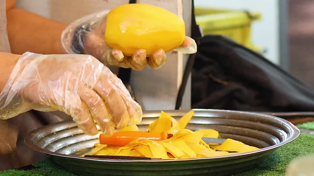 Un venditore di street food con guanti di plastica taglia con cura un mango giallo maturo a pezzetti