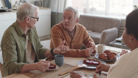 high angle medium close-up of two grey-haired caucasian men sitting at table at home, eating sweet cake, talking to unrecognizable people