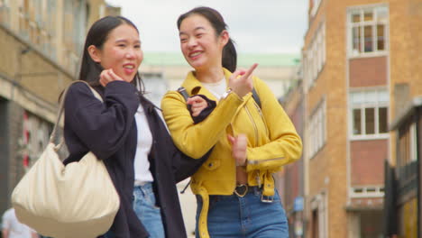 Two-Smiling-Young-Female-Friends-Walking-Arm-In-Arm-Along-City-Street-Together