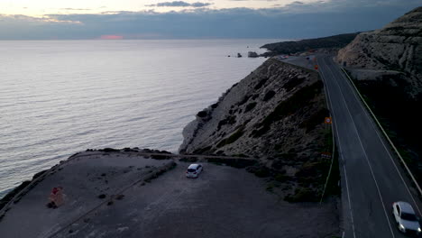 Aerial-view-at-dusk-of-a-coastal-road-near-Aphrodite's-Rock-in-Paphos,-with-a-parked-car-and-the-tranquil-sea-extending-into-the-horizon