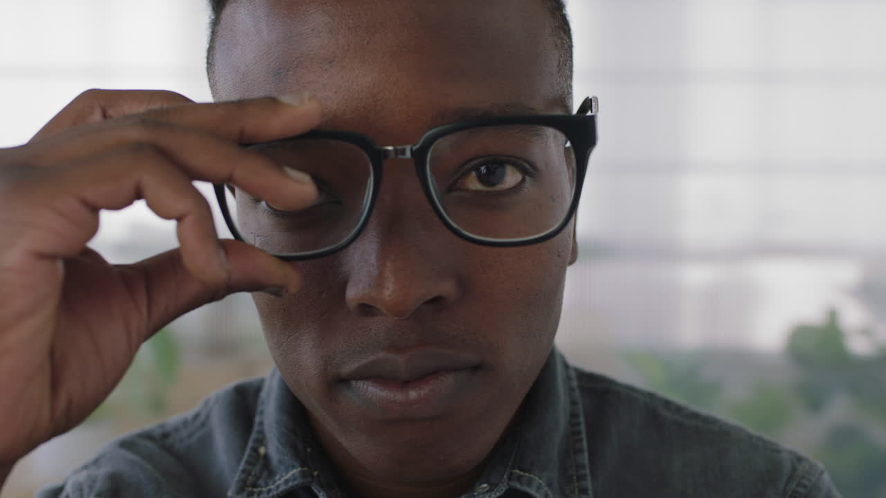 close up portrait of young male african american business student intern looking serious at camera puts on glasses smiling cheerful