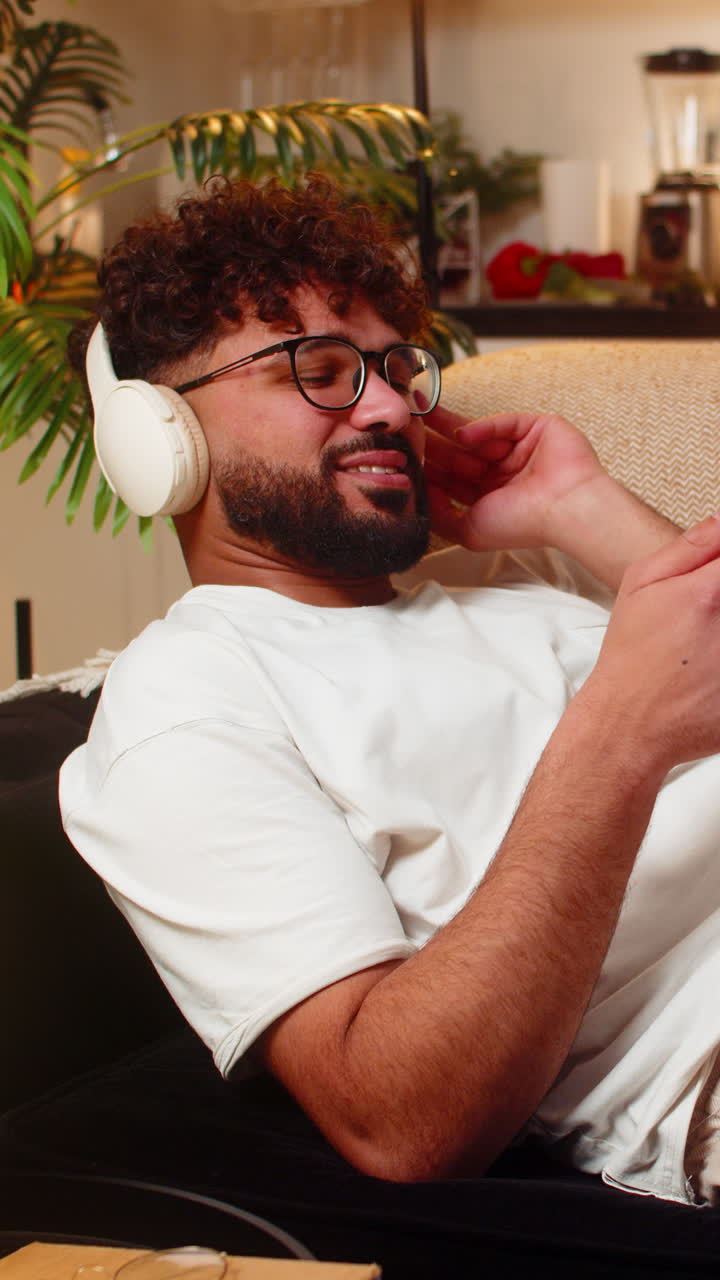 Uomo arabo con cuffie wireless ascolta musica e chatta su smartphone di notte, rilassato in un ambiente interno
