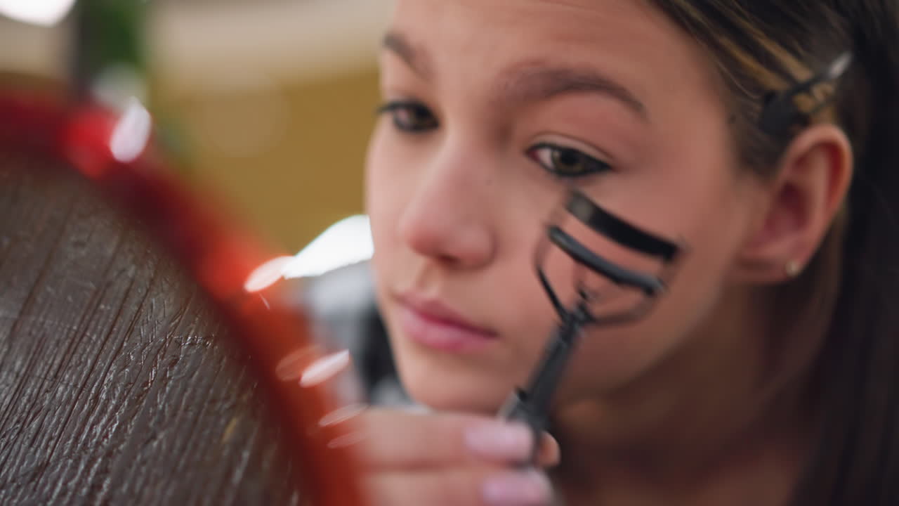 toma de primer plano de una chica usando un enroscador de pestañas para arreglar las pestañas mientras sostiene la herramienta cerca de su ojo con un reflejo borroso en el espejo que muestra un momento de aseo preciso para la mejora de las pestañas