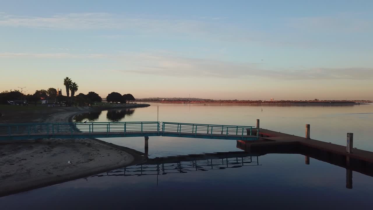 Drone che si allontana sull'acqua a Mission Bay, rivelando un molo per barche all'alba con una luce mattutina soffusa e un'atmosfera costiera.