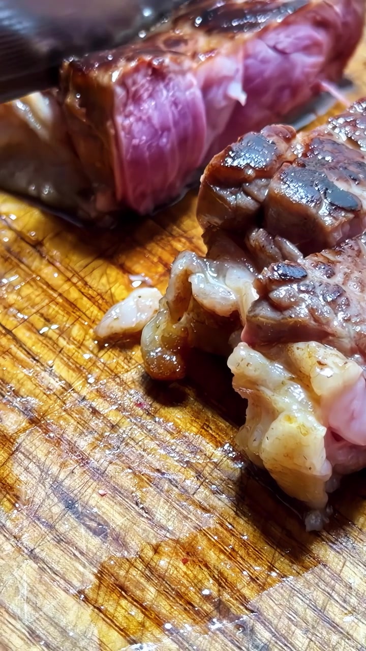 Una mano taglia una bistecca al sangue perfettamente cotta con marezzatura visibile e crosta rosolata su un tagliere di legno. Video verticale, ideale per i social media