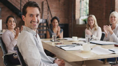 Portrait-of-a-confident-young-businessman--at-boardroom-table.-The-diverse-team-can-be-seen-chatting-together-in-the-background.-In-slow-motion
