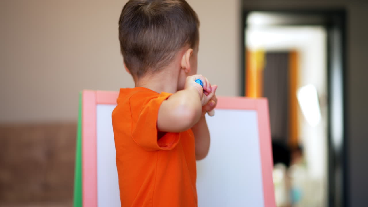 Lovely toddler in orange t-shirt comes up to a whiteboard. Baby boy takes and opens a blue marker.
